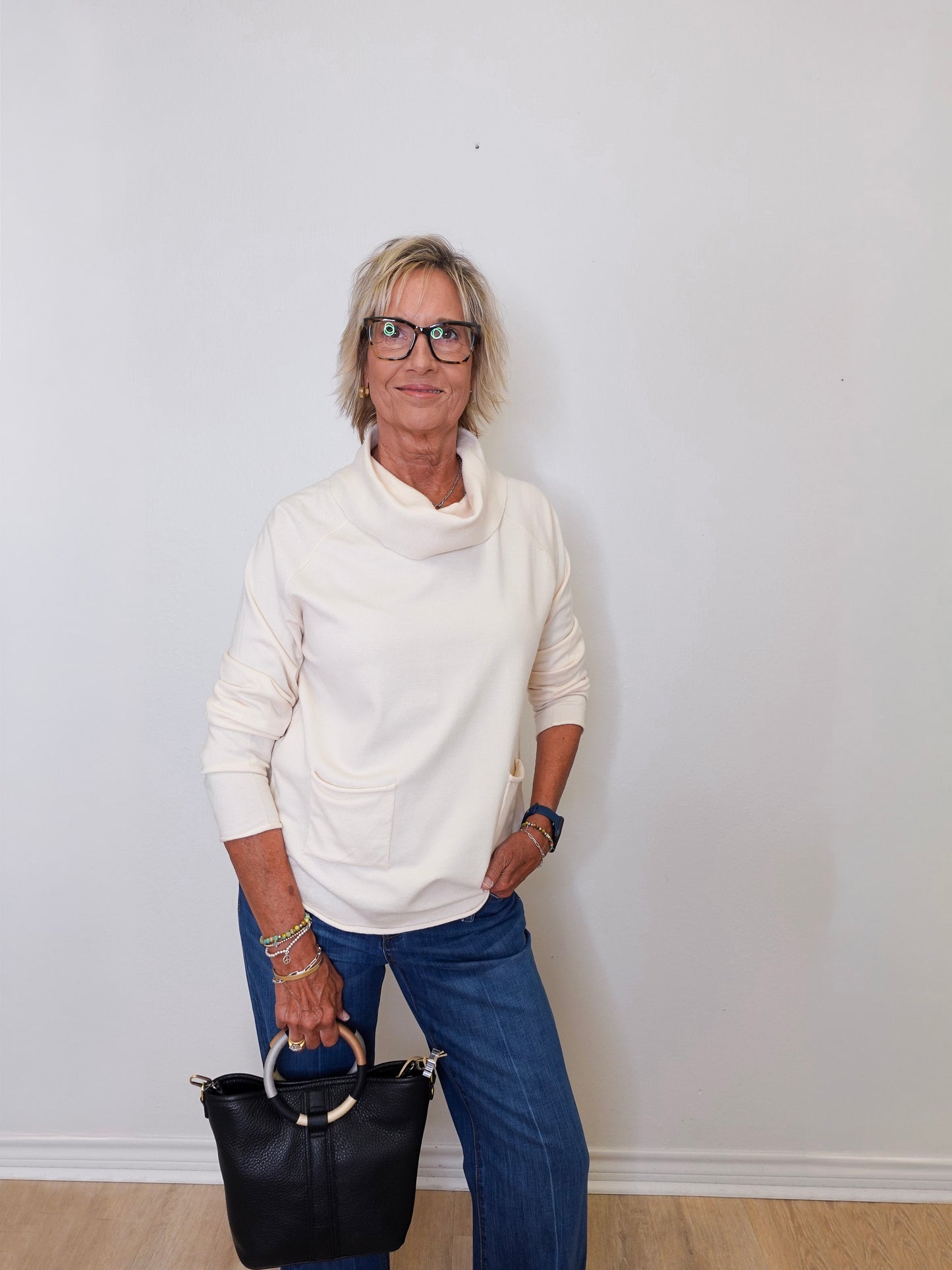A woman wearing a cowl neck sweater in cream with blue pants against a white wall.