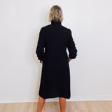 Person wearing a black coat standing against a white wall.