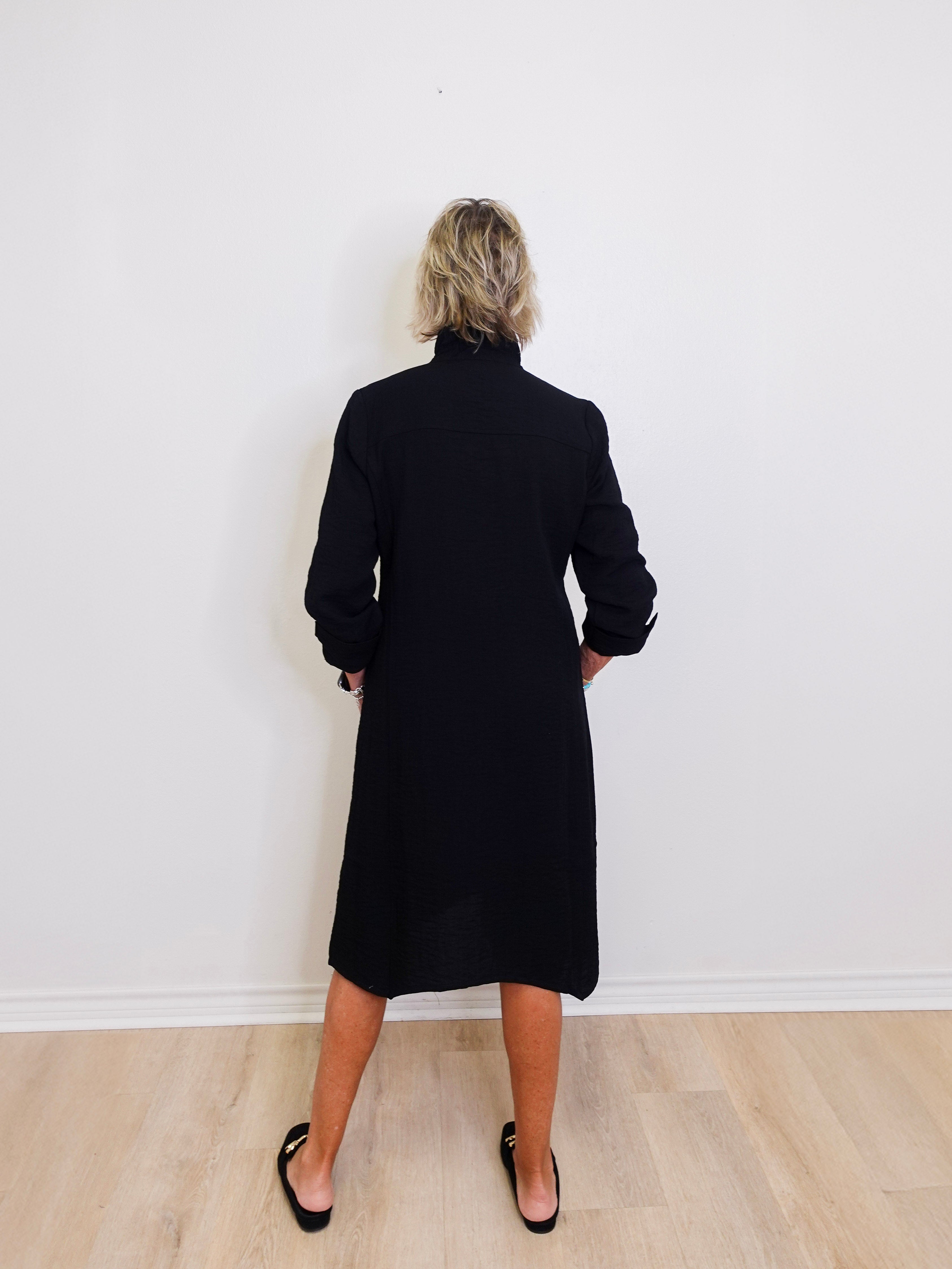 Person wearing a black coat standing against a white wall.