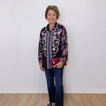 Woman wearing a patterned jacket and jeans standing against a white wall.