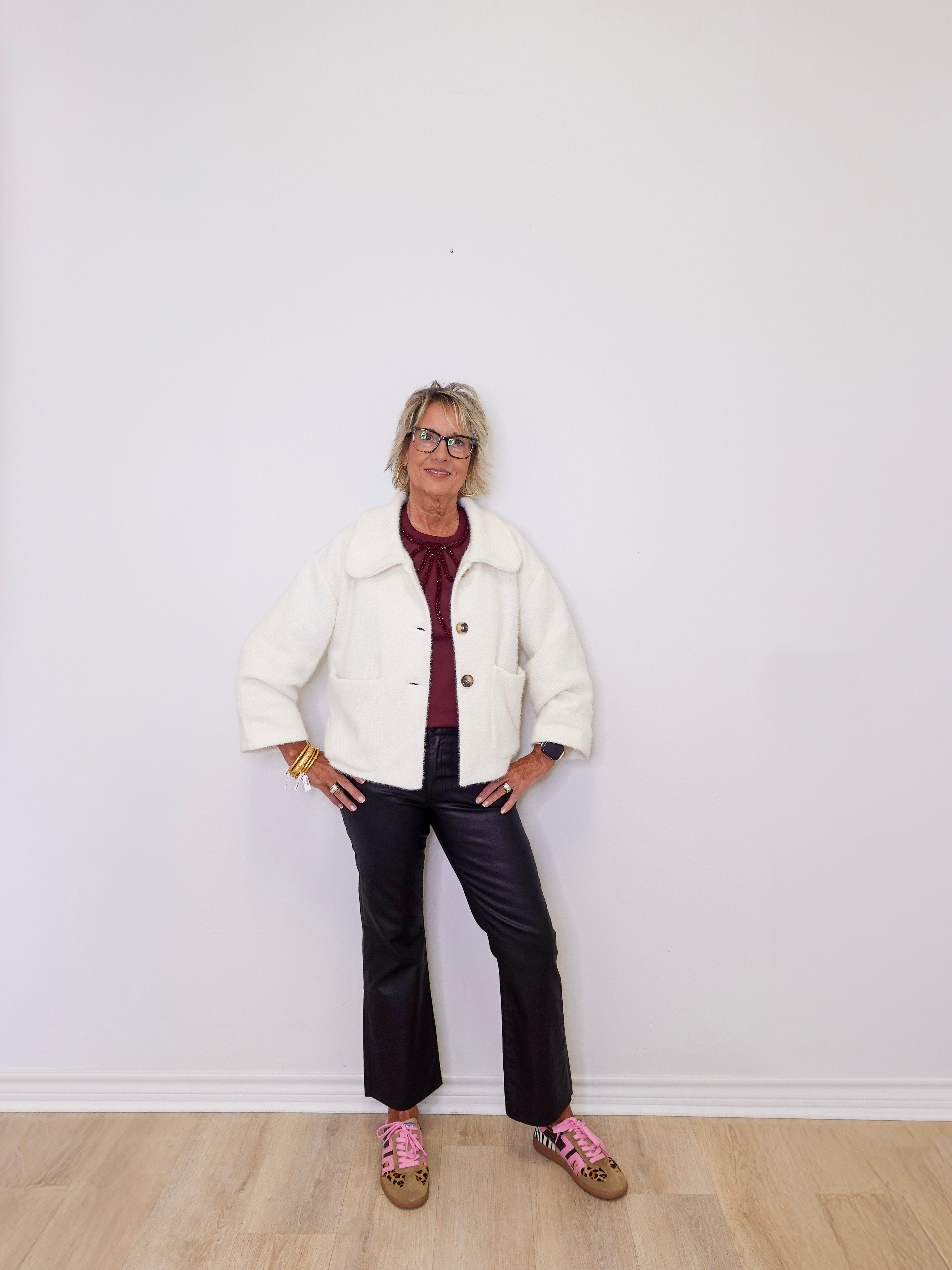 Person wearing a white jacket, red shirt, black pants, and pink shoes against a white wall.