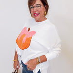 Woman wearing a white sweater with a colorful design, holding a brown handbag against a plain background.