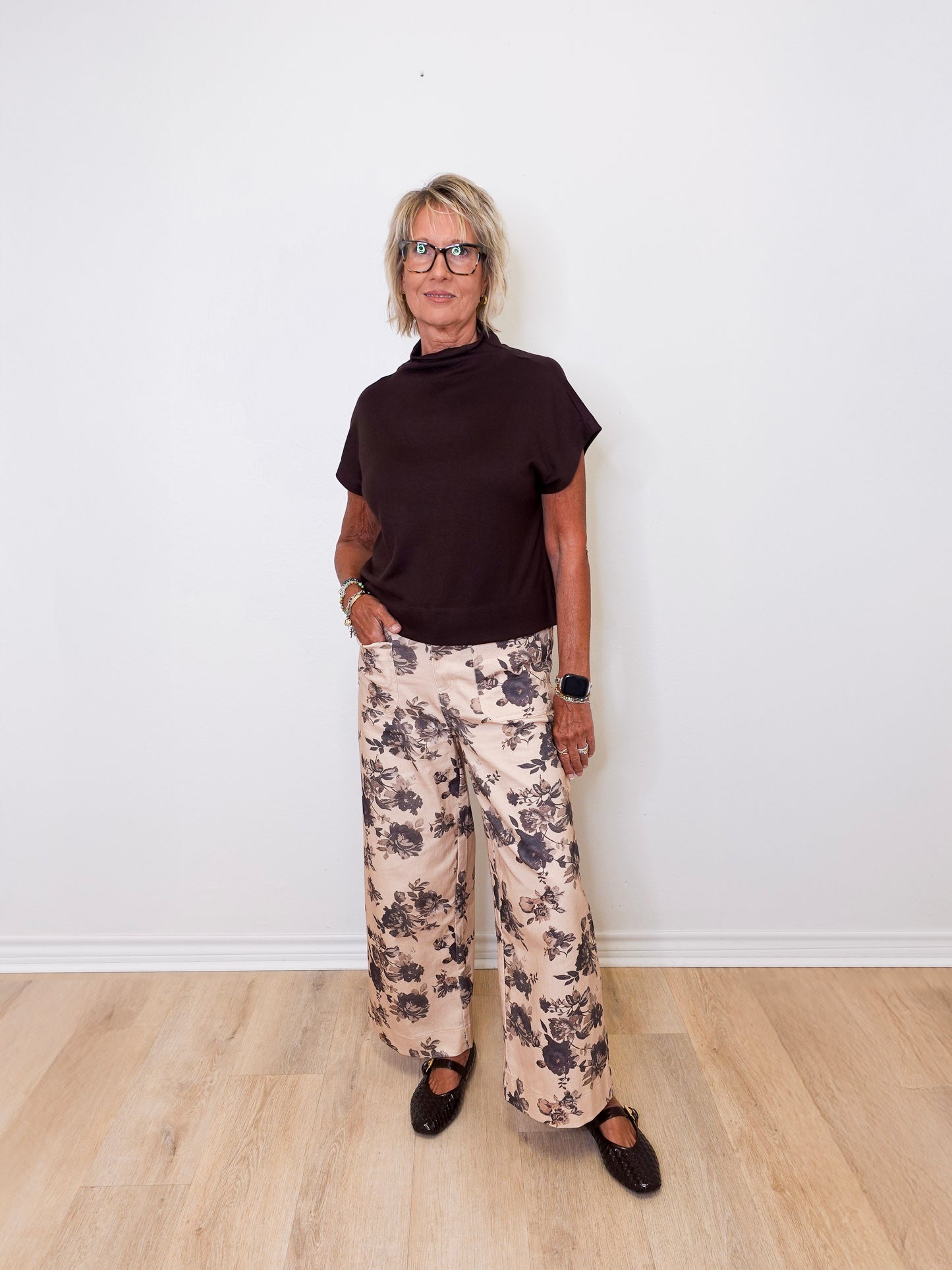 Person wearing a black top and floral pants standing against a white wall.