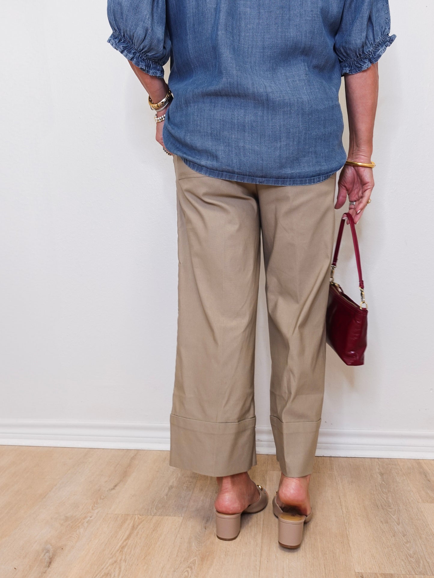 Person wearing a blue shirt and beige pants holding a red handbag against a white wall.