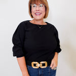 Woman wearing a black top and blue jeans with a gold belt against a white background