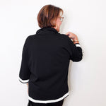 Person wearing a black jacket with white trim against a white wall