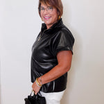 Woman wearing a black leather jacket and white pants, holding a black handbag against a white wall.