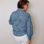 Person wearing a blue embroidered denim jacket and white shorts against a plain background