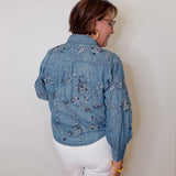 Person wearing a blue embroidered denim jacket and white shorts against a plain background