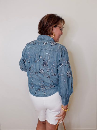Person wearing a blue embroidered denim jacket and white shorts against a plain background