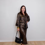 Woman wearing a leopard print outfit holding a black handbag against a white wall.