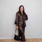 Woman wearing a leopard print outfit holding a black handbag against a white wall.