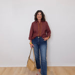 Woman wearing a plaid shirt and jeans, holding a beige handbag against a white wall.
