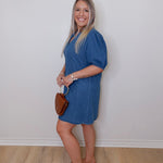 Woman wearing a blue dress holding a brown handbag against a white wall.