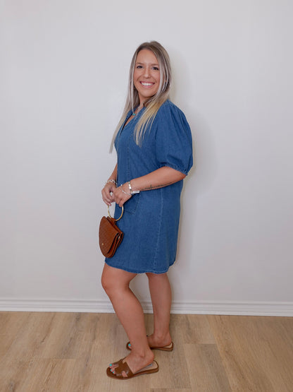 Woman wearing a blue dress holding a brown handbag against a white wall.