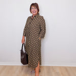 Woman wearing a patterned dress holding a handbag against a white wall.