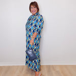 Woman wearing a blue patterned dress holding a handbag against a white wall.