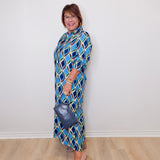 Woman wearing a blue patterned dress holding a handbag against a white wall.