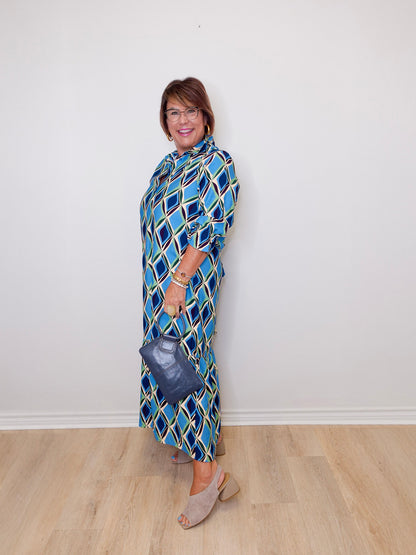 Woman wearing a blue patterned dress holding a handbag against a white wall.