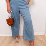Person wearing blue jeans and holding a brown handbag against a white wall.
