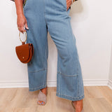Person wearing blue jeans and holding a brown handbag against a white wall.