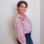 Woman wearing a patterned blouse with floral embroidery and dark jeans against a white wall.