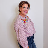 Woman wearing a patterned blouse with floral embroidery and dark jeans against a white wall.