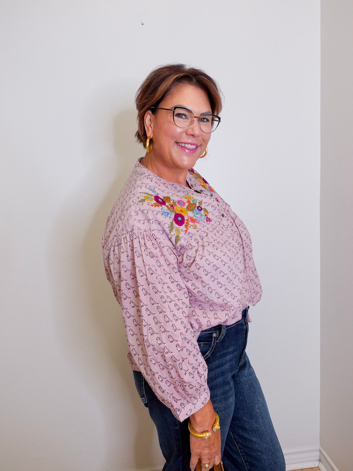 Woman wearing a patterned blouse with floral embroidery and dark jeans against a white wall.