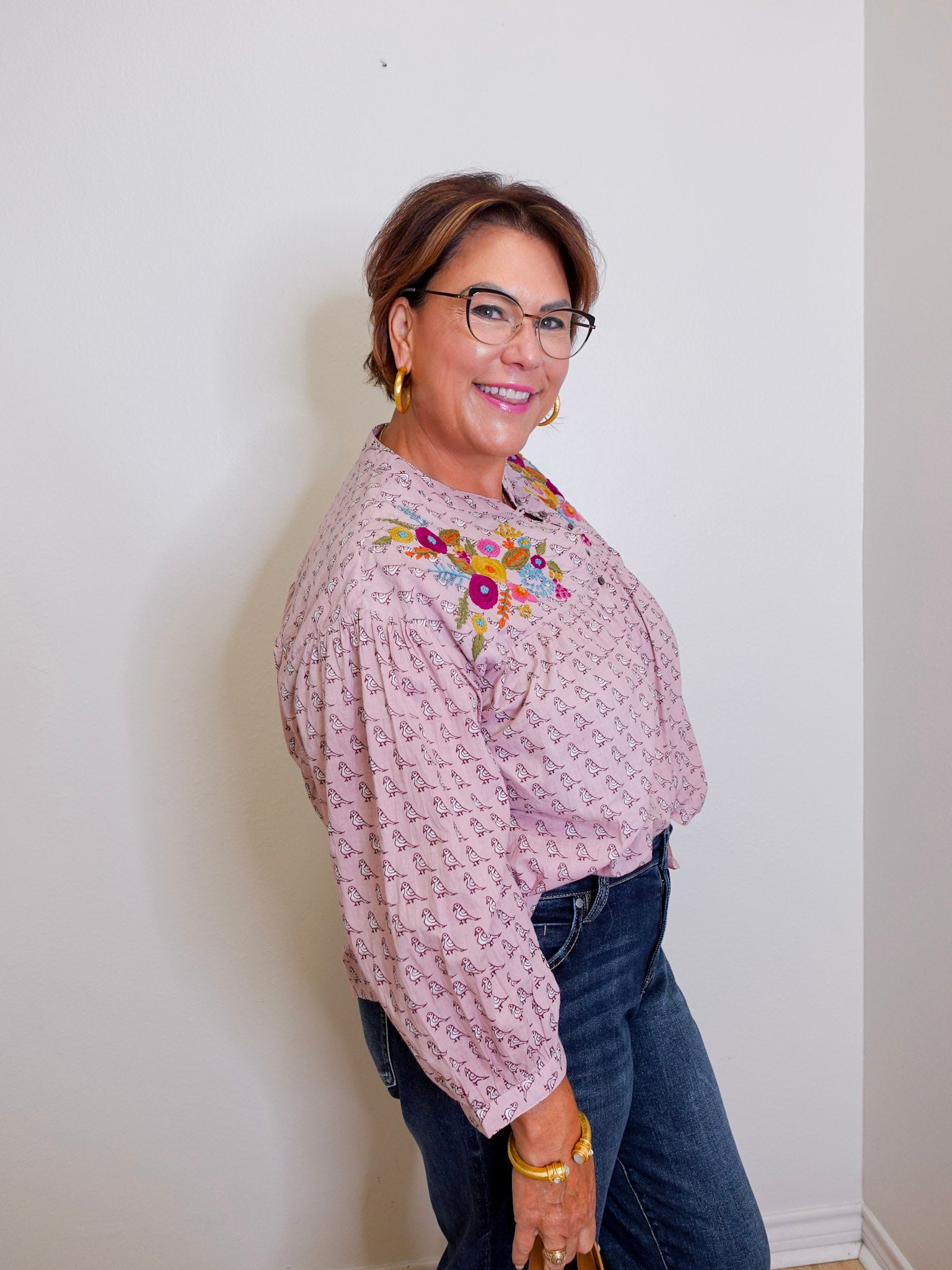 Woman wearing a patterned blouse with floral embroidery and dark jeans against a white wall.