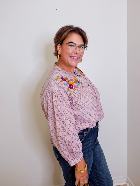 Woman wearing a patterned blouse with floral embroidery and dark jeans against a white wall.