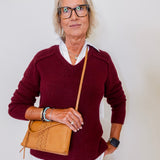 Woman wearing a maroon sweater and holding a tan handbag against a plain background