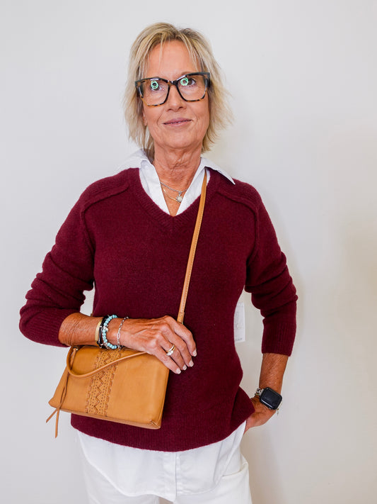 Woman wearing a maroon sweater and holding a tan handbag against a plain background