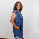 Woman wearing a blue dress with white embroidery against a white wall.