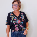 Woman wearing a black top with colorful floral embroidery against a white background