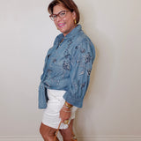 Woman wearing a blue embroidered shirt and white shorts against a plain wall.