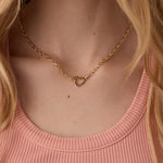Gold heart necklace worn by a person with blonde hair and a pink top.