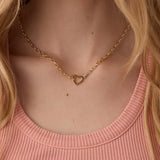 Gold heart necklace worn by a person with blonde hair and a pink top.