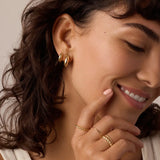 Woman wearing gold hoop earrings and rings with a neutral background