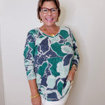 Woman wearing a patterned top and white pants against a plain background