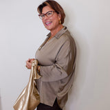 Woman wearing glasses and a beige shirt, holding a gold bag against a white wall.