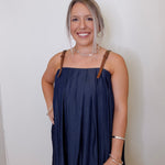 Woman wearing a navy blue dress with brown straps against a white wall.