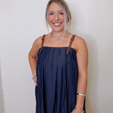 Woman wearing a navy blue dress with brown straps against a white wall.