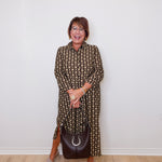 Woman wearing a patterned dress and holding a handbag against a white wall.