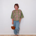 Woman wearing a patterned top and jeans, holding a handbag against a white wall.