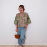 Woman wearing a patterned top and jeans, holding a handbag against a white wall.