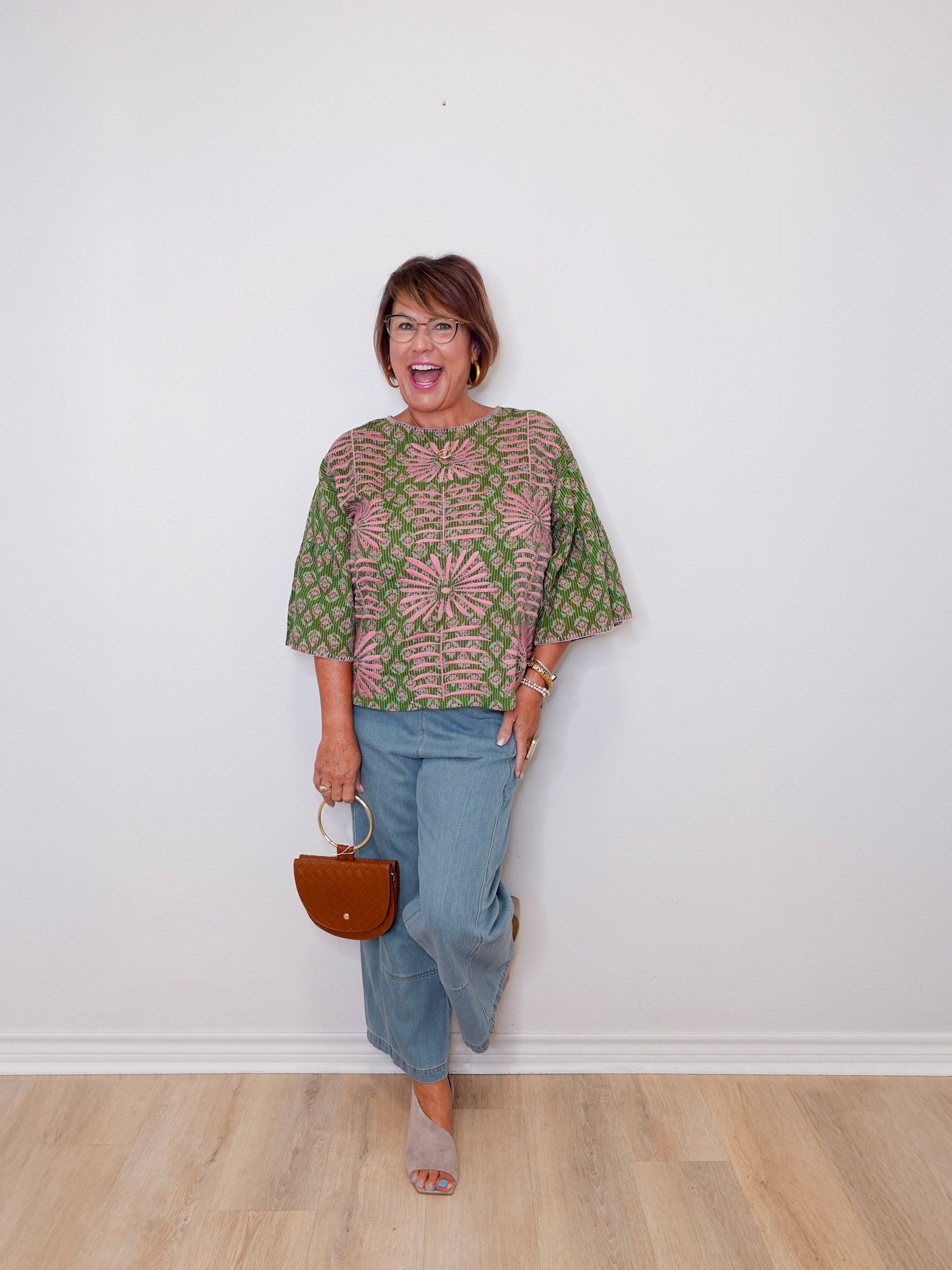Woman wearing a patterned top and jeans, holding a handbag against a white wall.