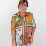 A woman with short brown hair and glasses wearing a colorful, patterned button-up shirt with three-quarter sleeves and light-wash jeans, standing against a plain white background.