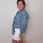 Woman wearing a blue floral shirt and white shorts, standing against a plain wall.