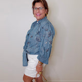 Woman wearing a blue floral shirt and white shorts, standing against a plain wall.