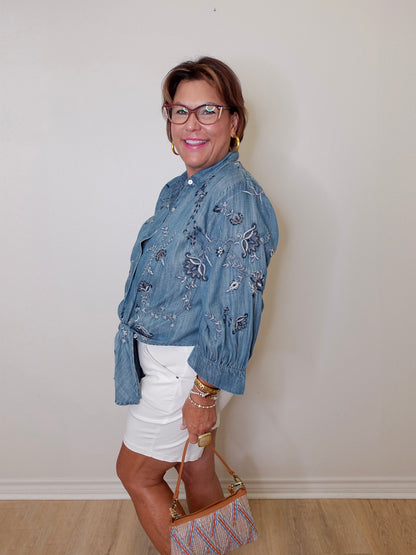 Woman wearing a blue floral shirt and white shorts, standing against a plain wall.